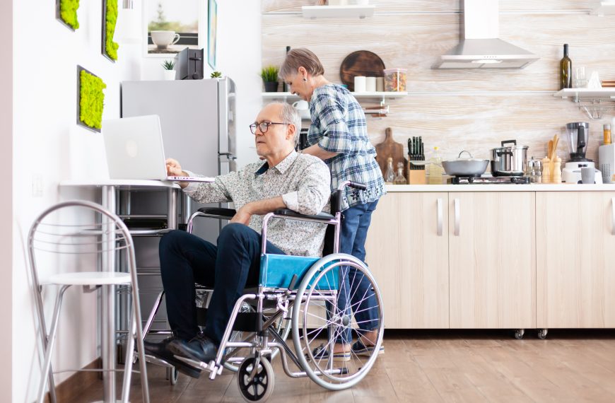 elderly man in a wheelchair typing on a laptop in a kitchen